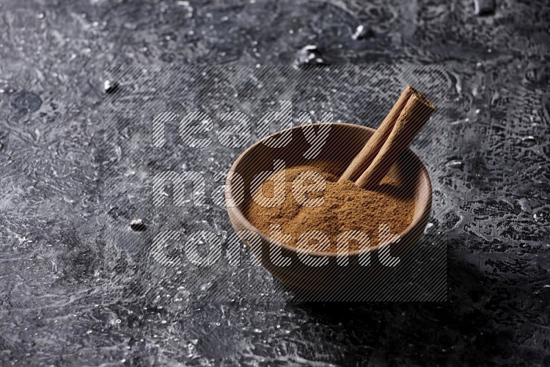 Wooden bowl full of cinnamon powder and a cinnamon stick on a textured black background