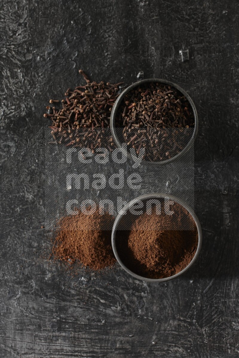 2 Black pottery bowls full of cloves and the other full of cloves powder on textured black flooring