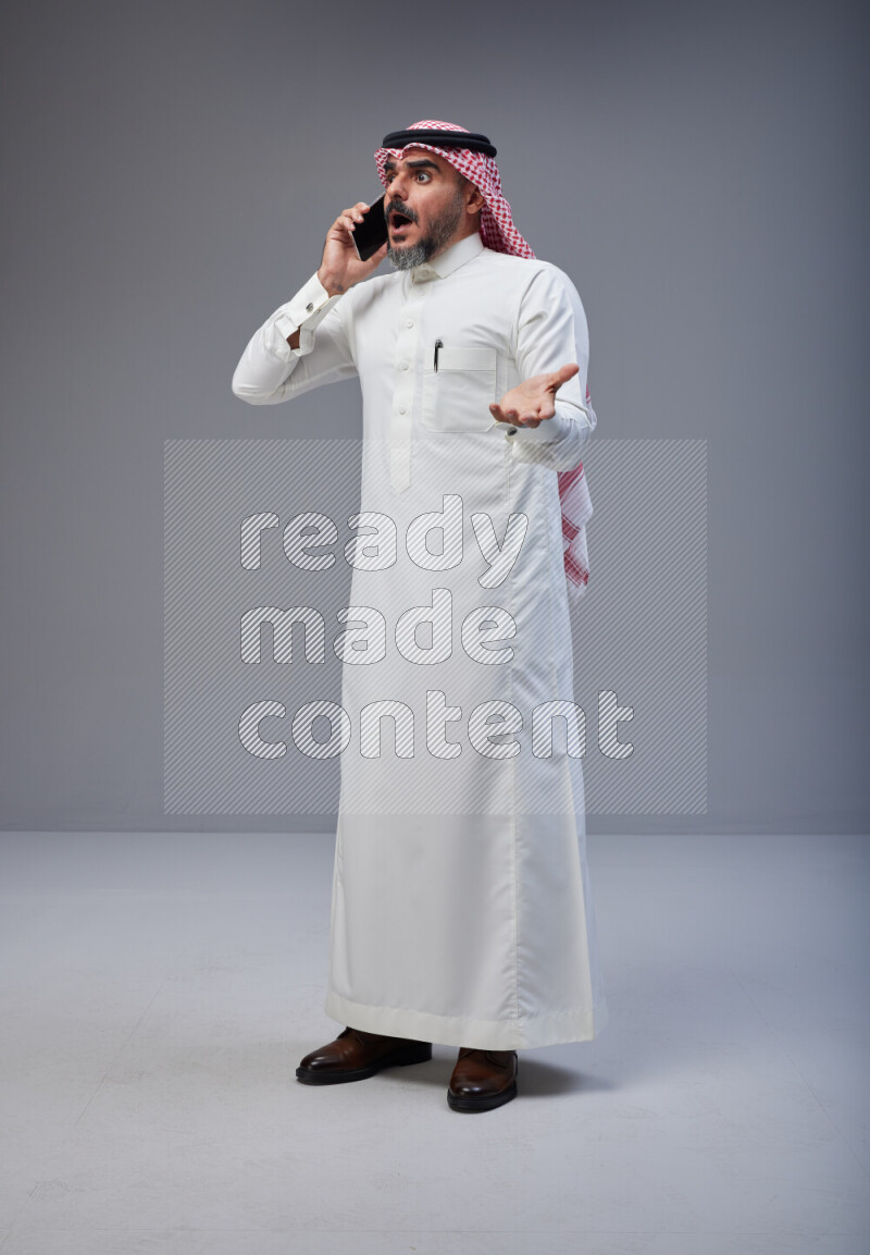 Saudi man Wearing Thob and red Shomag standing talking on phone on Gray background