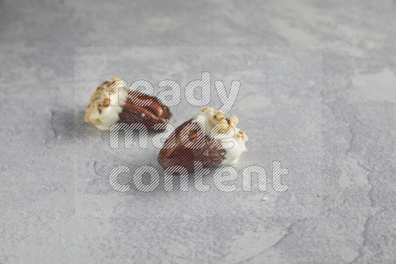 Two pecan stuffed date covered with white chocolate and chopped walnuts on alight grey background