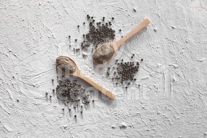 2 wooden spoons full of black pepper powder and black pepper beads spread on textured white background