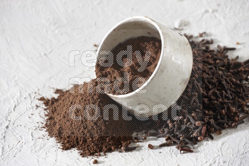 وعاء بيج من السيراميك ممتلئ ببودرة القرنفل وبعض بودرة القرنفل سقطت بخارجه على أرضية بيضاء