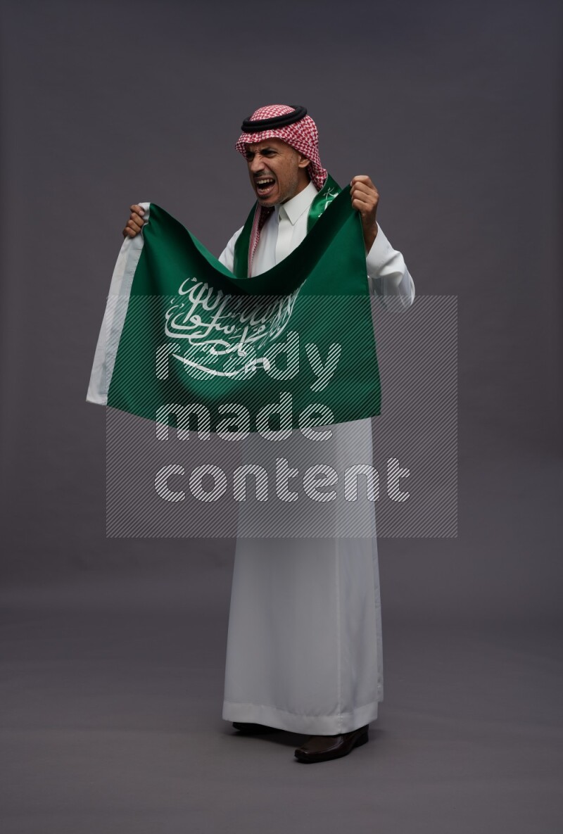 Saudi man wearing thob and shomag standing holding Saudi flag on gray background