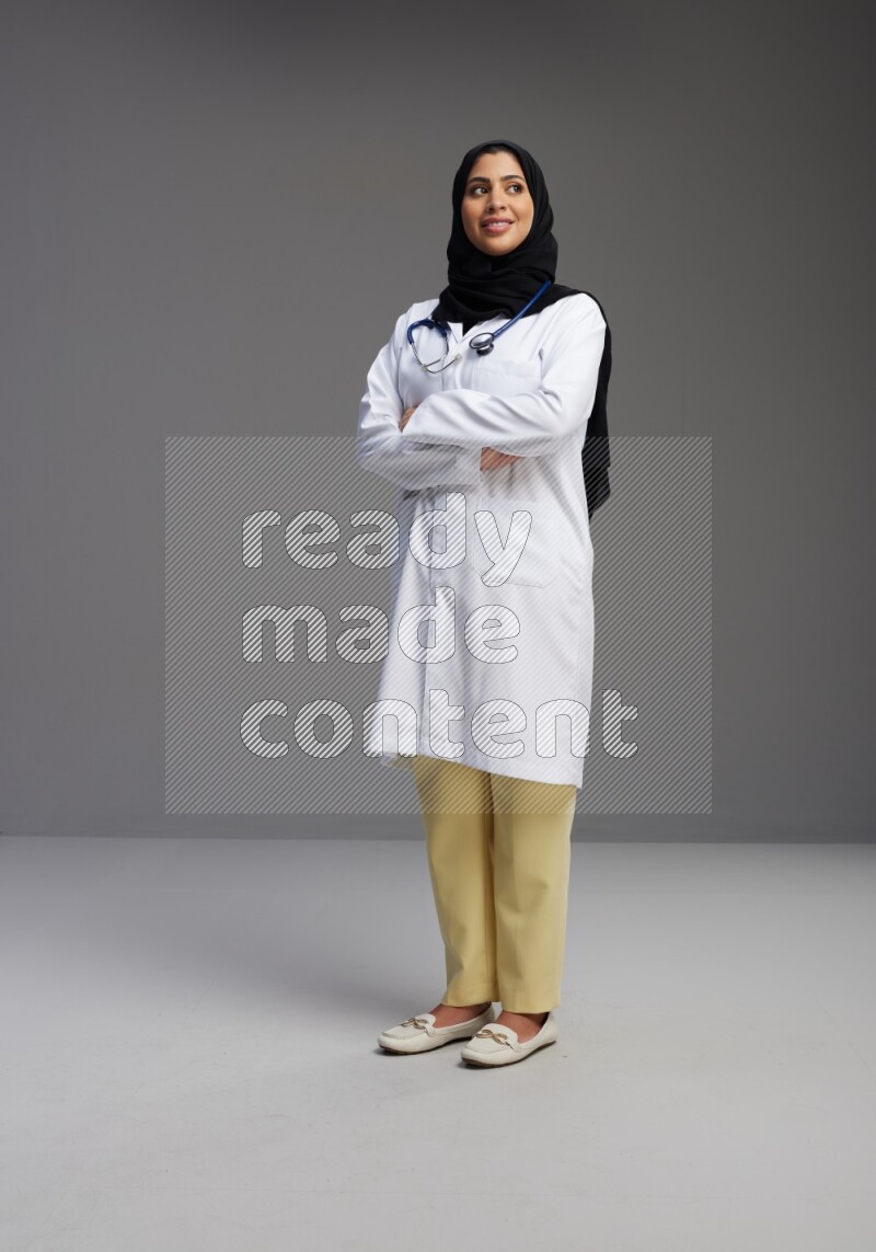 Saudi woman wearing lab coat with stethoscope standing with crossed arms on Gray background