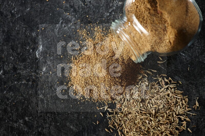 A flipped glass spice jar full of cumin powder with spilled powder and cumin seeds on a textured black flooring