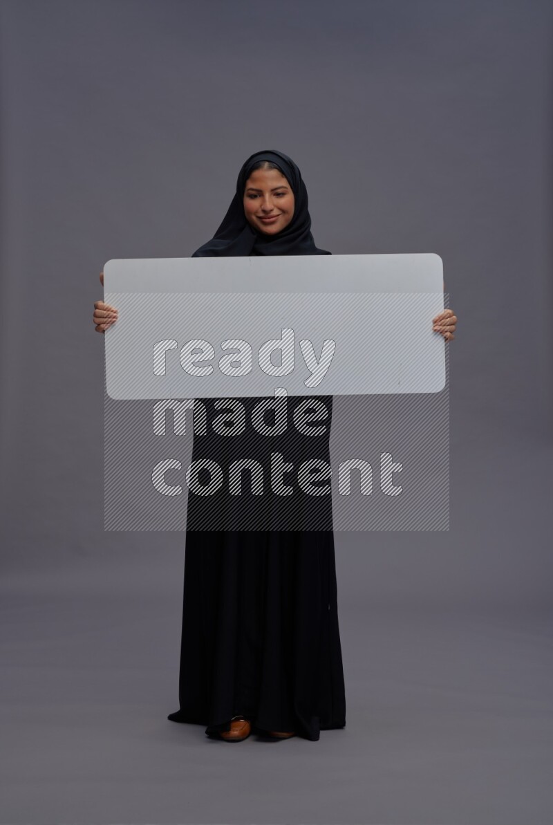 Saudi woman wearing Abaya standing holding white board on gray background