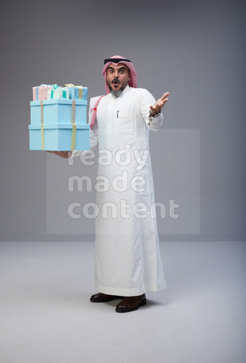 Saudi man Wearing Thob and red Shomag standing holding gift box on Gray background