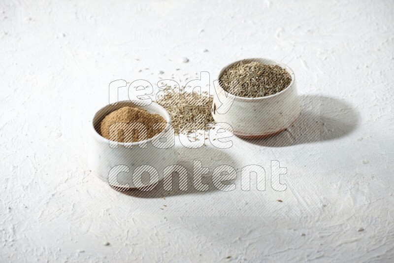 2 beige bowls full of cumin seeds and powder with spilled powder and seeds on textured white flooring