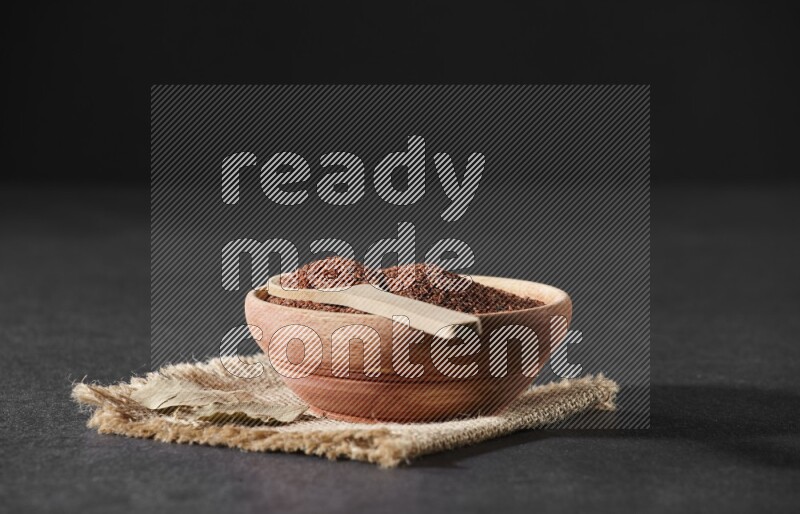 A wooden bowl full of garden cress seeds with wooden spoon full of the seeds on it on burlap fabric on a black flooring