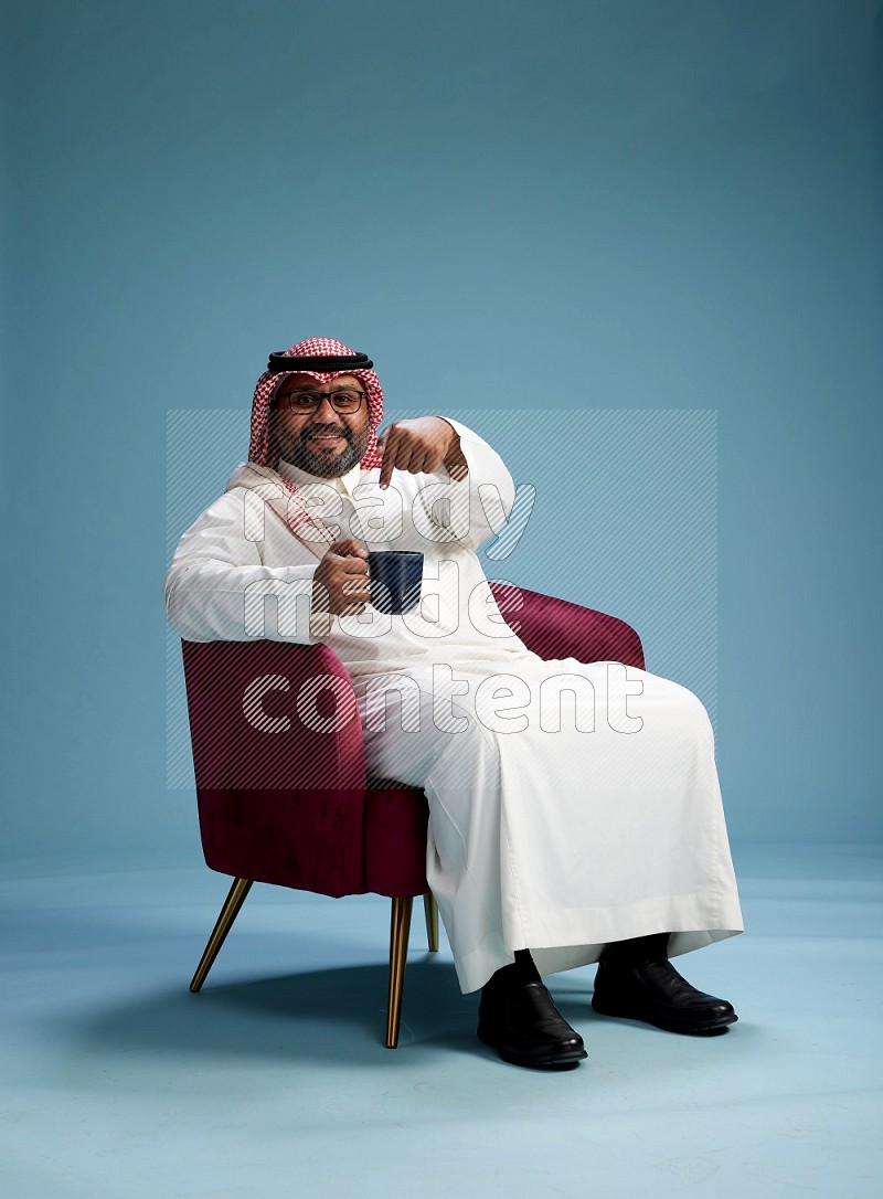 Saudi Man with shimag sitting on chair drinking coffee on blue background