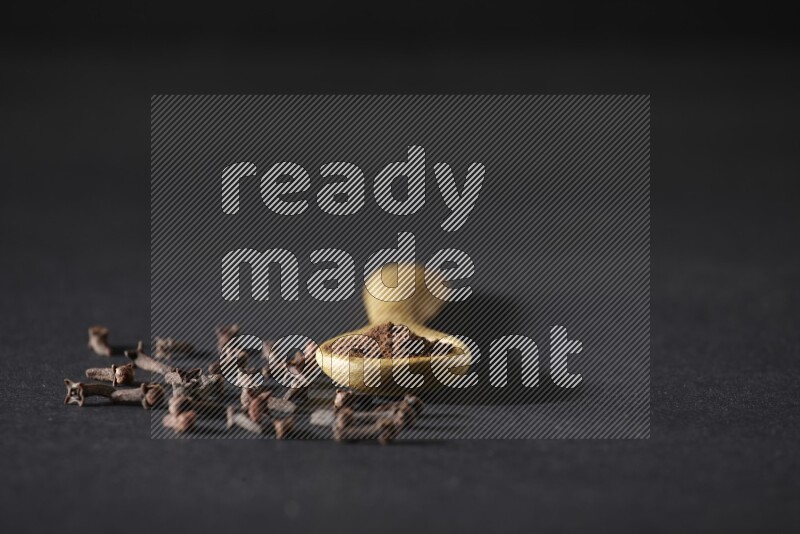 ملعقة خشبية ممتلئة ببودرة القرنفل مع حبوب القرنفل المتناثرة على خلفية سوداء