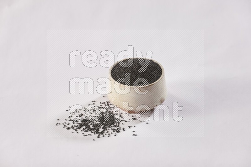 A beige pottery bowl full of black seeds and more seeds spread on a white flooring