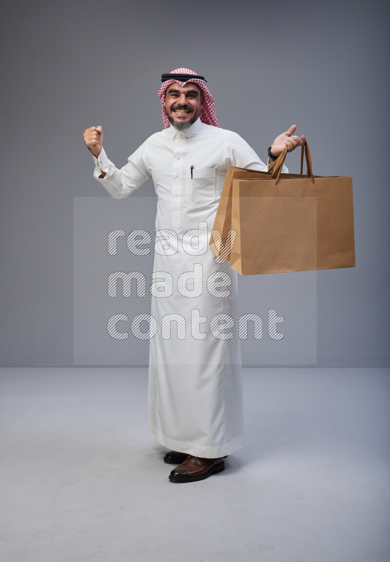 Saudi man Wearing Thob and red Shomag standing holding shopping bag on Gray background