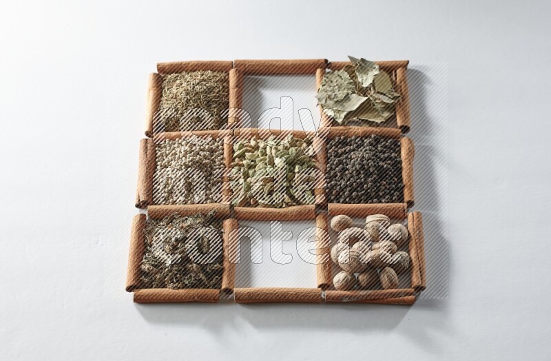 9 مربعات من أعواد القرفة ممتلئة بالهيل في المنتصف محاطة بجوزة الطيب والملح والريحان المجفف والفلفل الأبيض والكمون وبهار الفلفل الحلو وأوراق الغار والفلفل الأسود على أرضية بيضاء