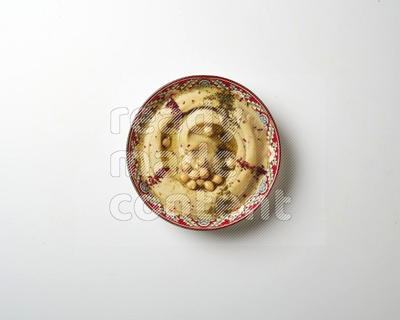 Hummus in a red plate with patterns garnished with zaatar & sumak on a white background