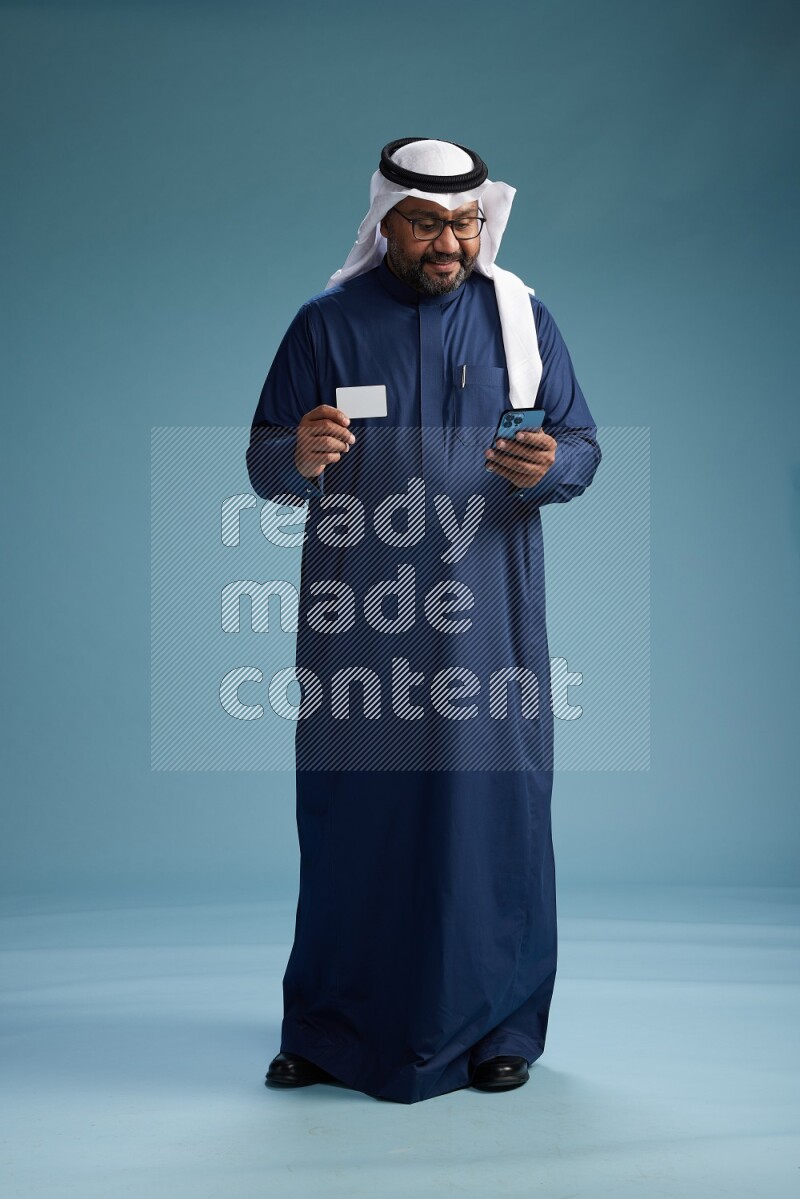 Saudi Man with shimag Standing holding ATM card while talking on phone on blue background
