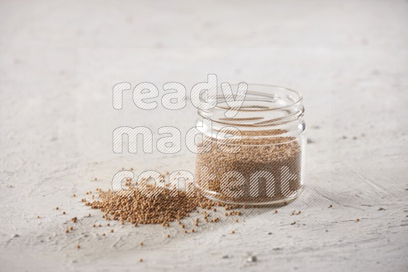 A glass jar full of mustard seeds and more seeds spread on a textured white flooring