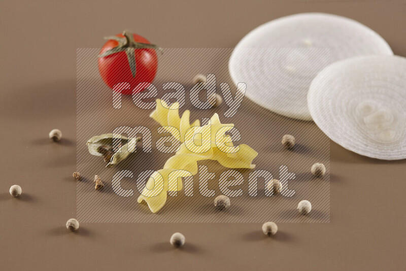 Raw pasta with different ingredients such as cherry tomatoes, garlic, onions, red chilis, black pepper, white pepper, bay laurel leaves, rosemary and cardamom on beige background