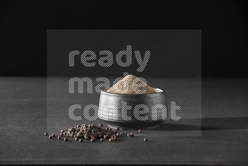 A black pottery bowl full of black pepper powder and black pepper beads on the floor on black flooring