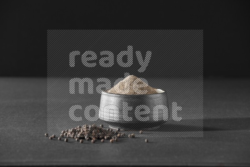 بودرة الفلفل الأسود في وعاء فخاري أسود مع حبوب الفلفل الأسود بجانبها علي خلفية سوداء