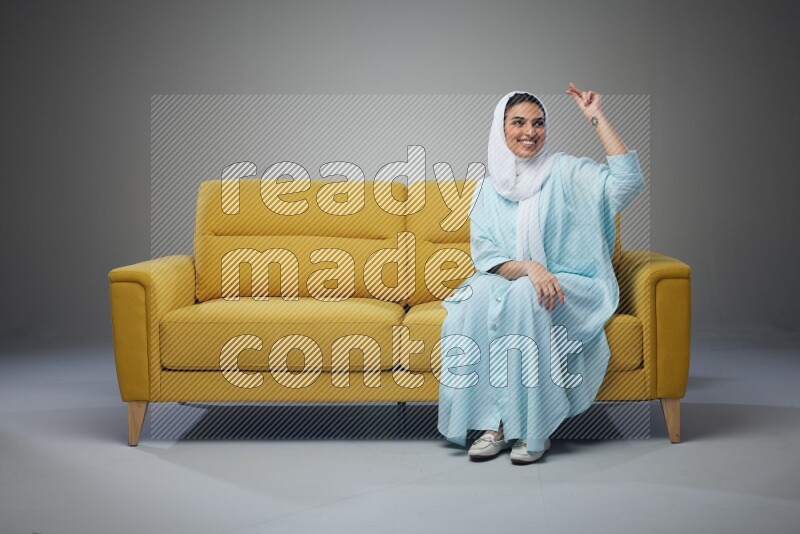 A Saudi woman wearing a light blue Abaya and white head scarf sitting on a yellow sofa and thinking and pointing up eye level on a grey background