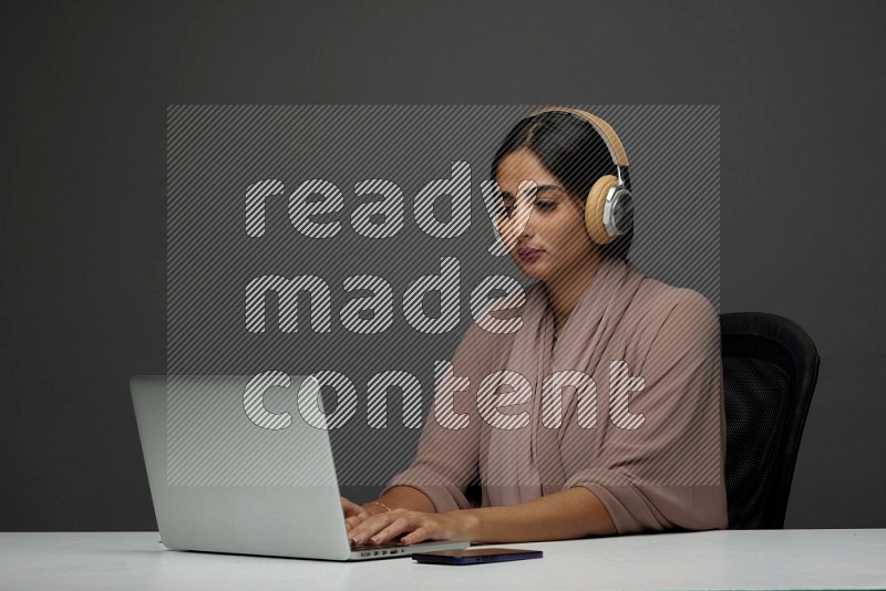 A Saudi woman Sitting on her desk Typing on her laptop wearing a headset  on a Gray Background wearing Brown Abaya