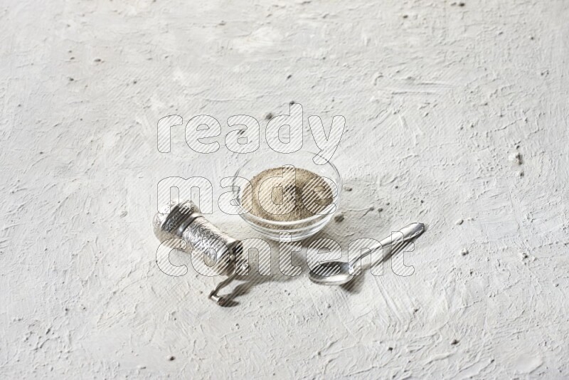 وعاء زجاجي ممتلئ ببودرة الفلفل الأبيض مع حبوب الفلفل الأبيض ومطحنة معدنية على أرضية بيضاء