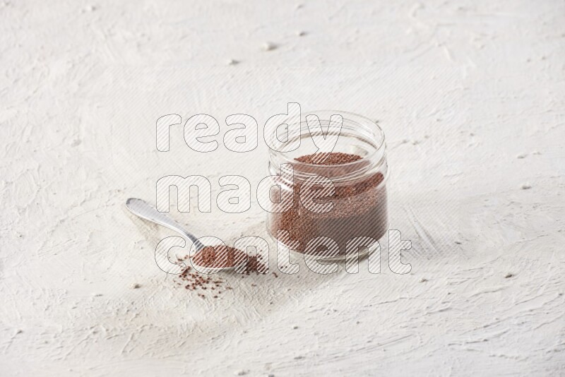 A glass jar and a metal spoon full of garden cress seeds on a textured white flooring