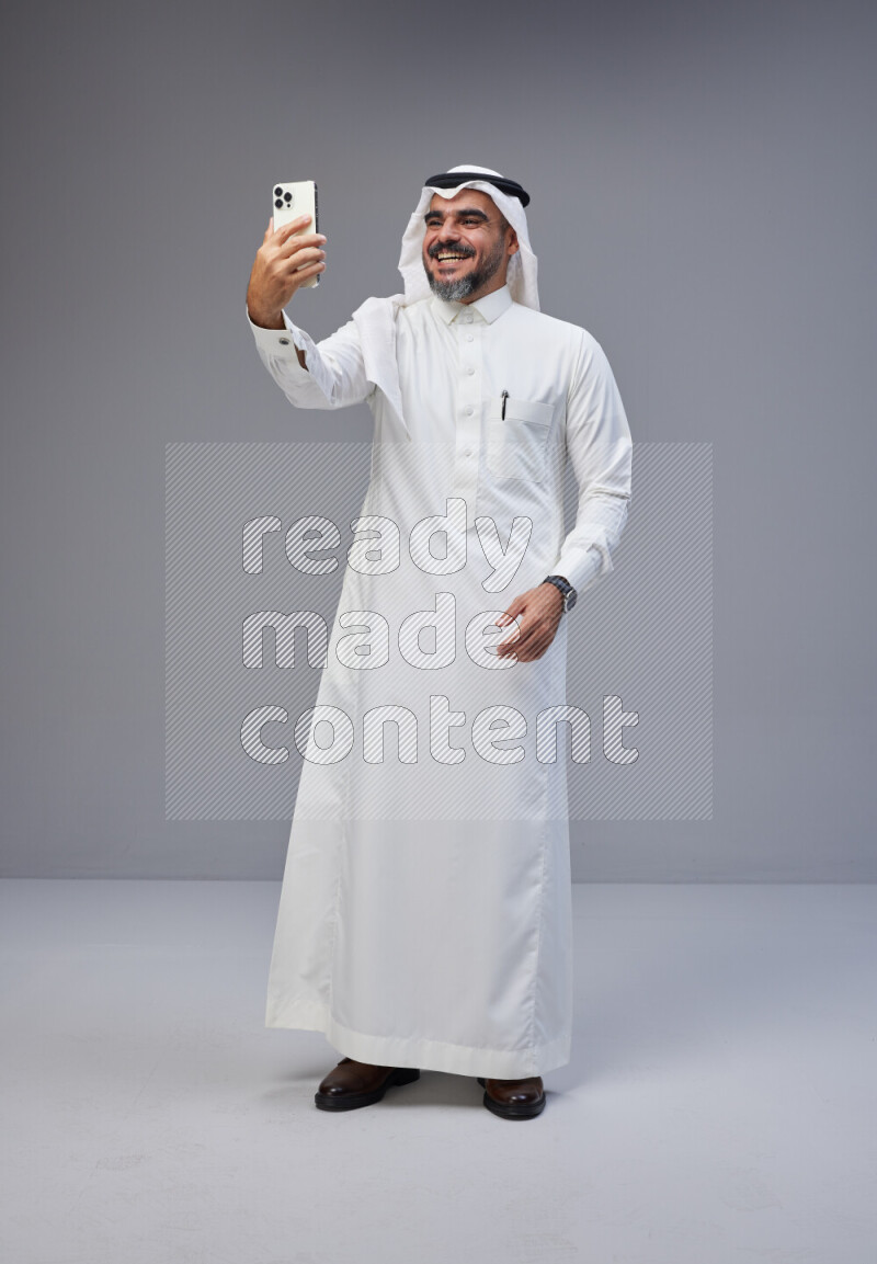 Saudi man Wearing Thob and white Shomag standing taking selfie on Gray background