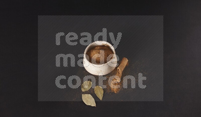 Cinnamon powder in a white pottery bowl and cinnamon sticks and laurel leaves on black background