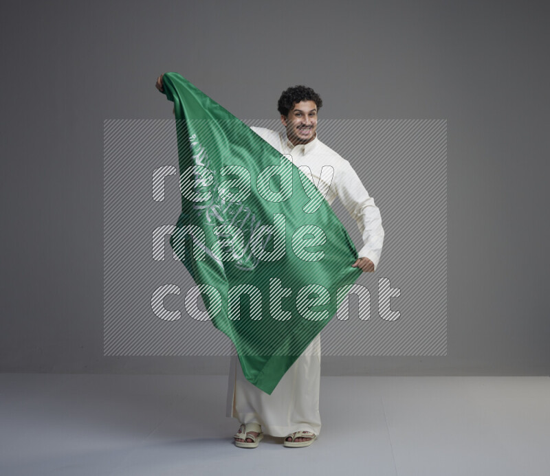 A saudi man standing wearing thob holding big saudi flag on gray background