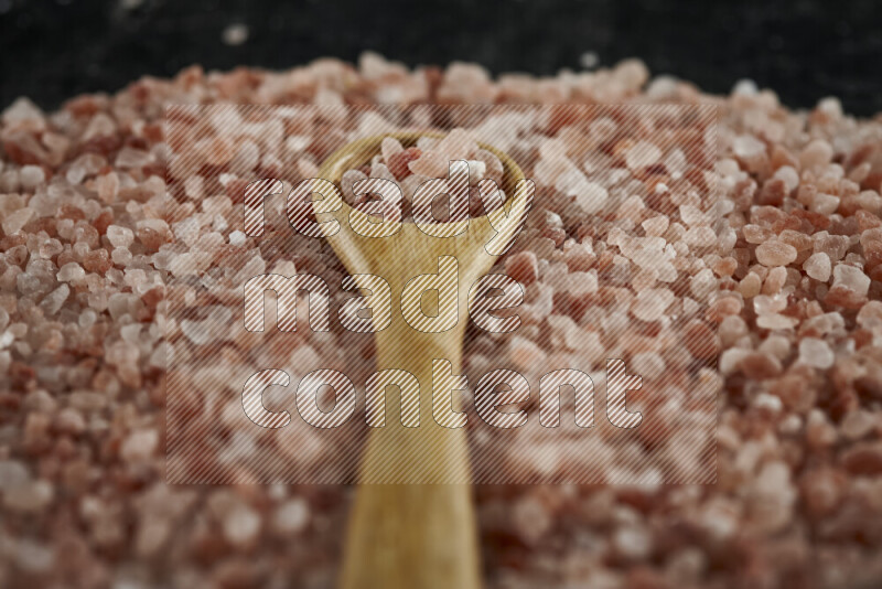 A wooden spoon full of coarse himalayan salt crystals on a bunch of the crystals on black background