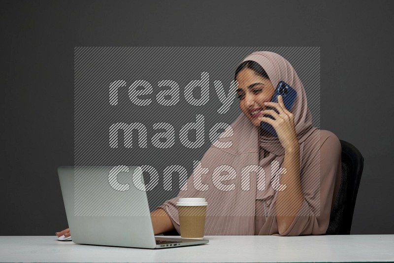 A Saudi woman Setting on her desk
 calling  on a Gray Background wearing Brown Abaya with Hijab