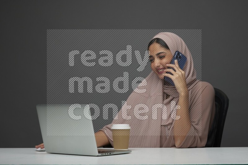 A Saudi woman Setting on her desk
 calling  on a Gray Background wearing Brown Abaya with Hijab
