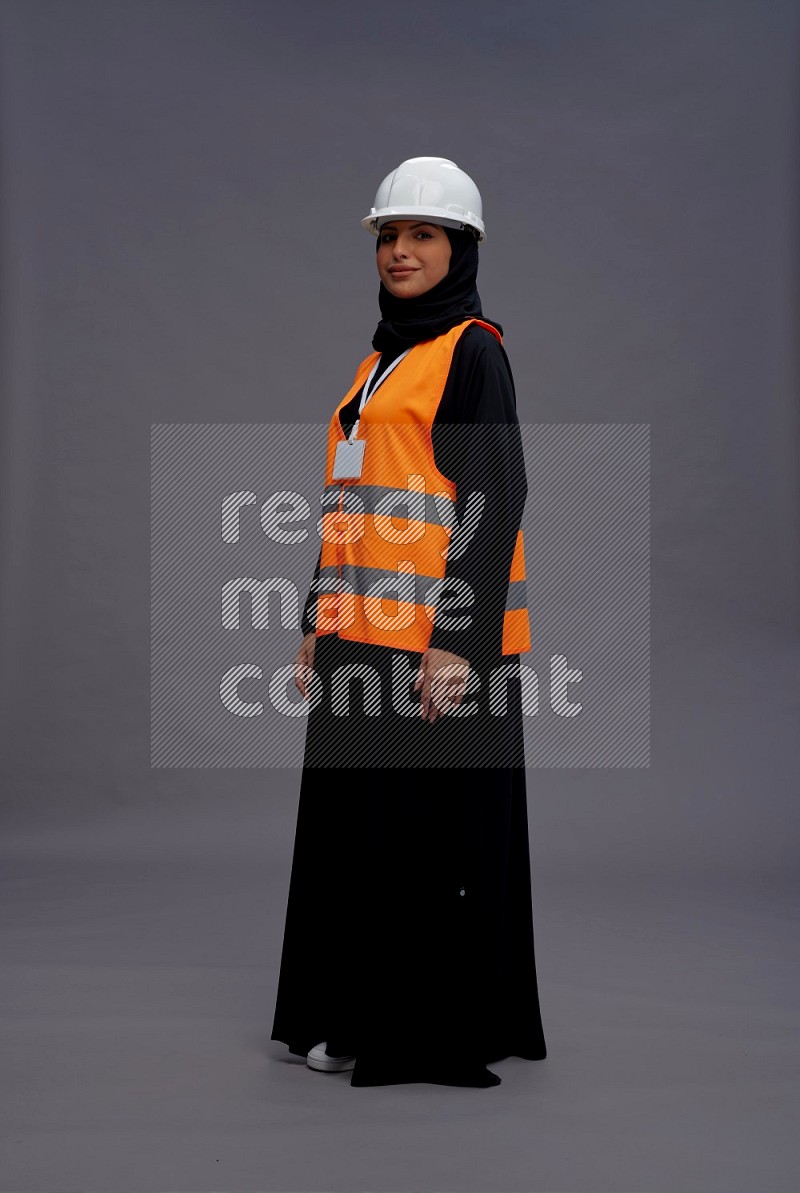 Saudi woman wearing Abaya with engineer vest with neck strap employee badge standing interacting with the camera on gray background