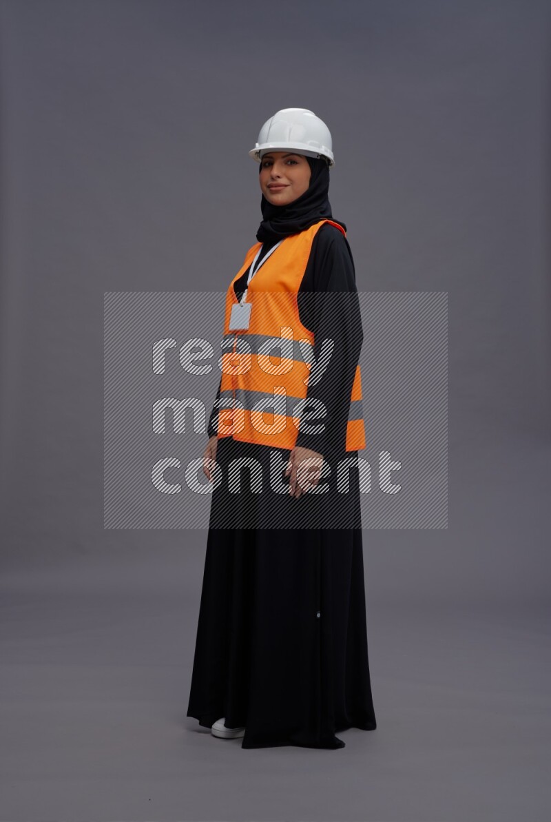 Saudi woman wearing Abaya with engineer vest with neck strap employee badge standing interacting with the camera on gray background