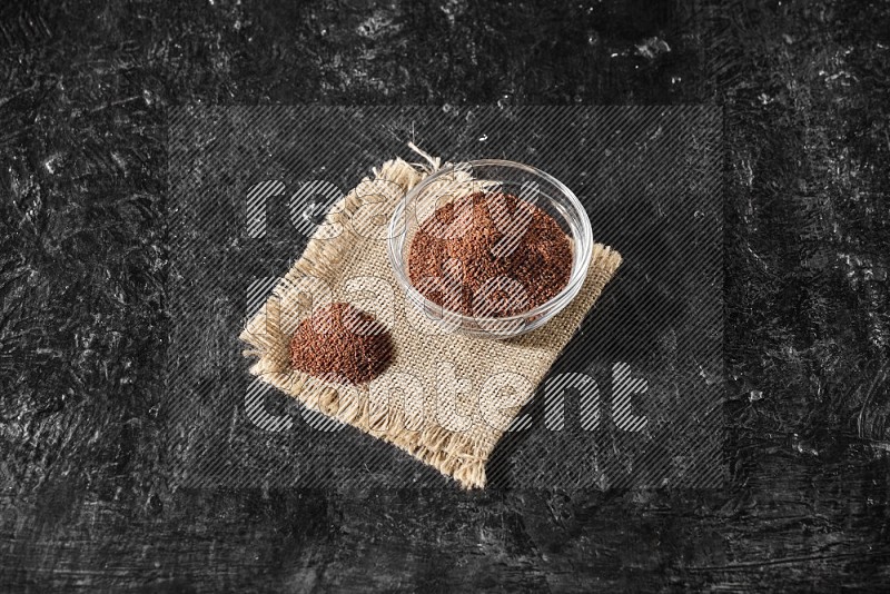 A glass bowl full of garden cress seeds with bunch of the seeds on burlap fabric on a textured black flooring