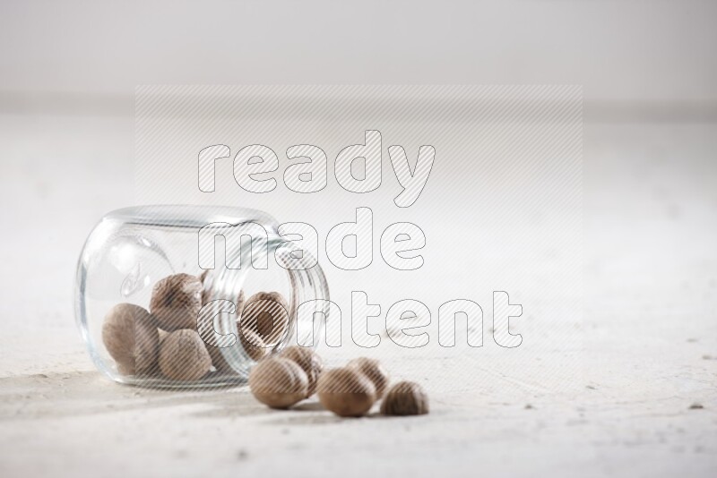 وعاء زجاجي ممتلئ بجوزة الطيب مقلوب وبعض الحبوب متناثرة على خلفية بيضاء