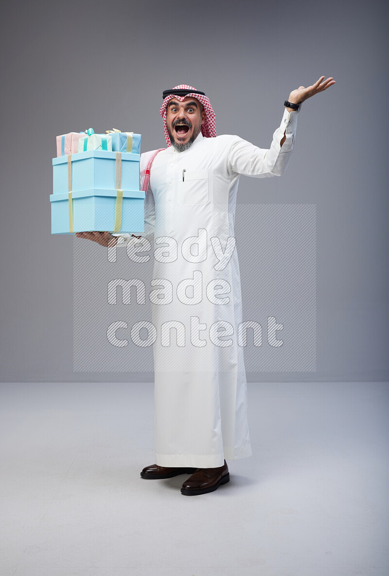 Saudi man Wearing Thob and red Shomag standing holding gift box on Gray background