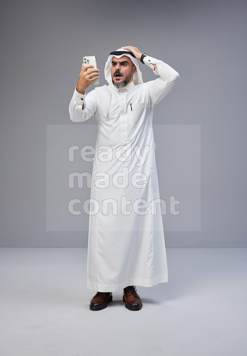 Saudi man Wearing Thob and white Shomag standing texting on phone on Gray background