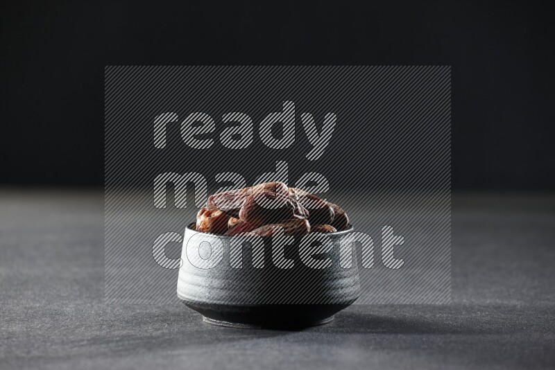 A black pottery bowl full of dried dates on a black background in different angles