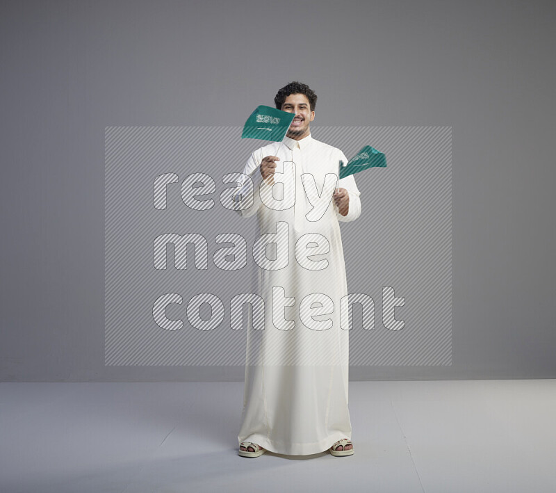 A Saudi man standing wearing thob holding small Saudi flag on gray background