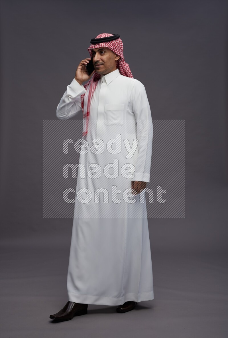 Saudi man wearing thob and shomag standing talking on phone on gray background