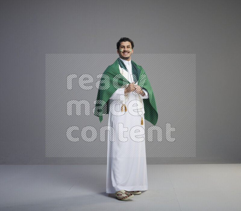 A Saudi man standing wearing thob and saudi flag scarf with face painting wrapping big Saudi flag on gray background