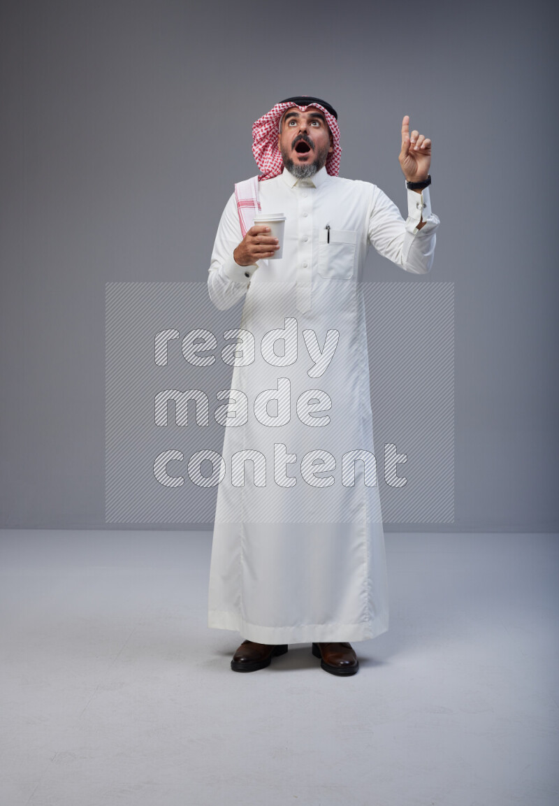 Saudi man Wearing Thob and red Shomag standing holding paper cup on Gray background