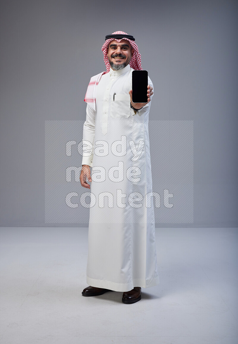 Saudi man Wearing Thob and red Shomag standing showing phone to camera on Gray background