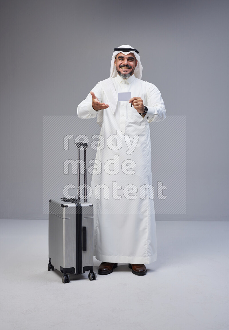 Saudi man wearing Thob and white Shomag standing holding Travel bag and ATM card on Gray background