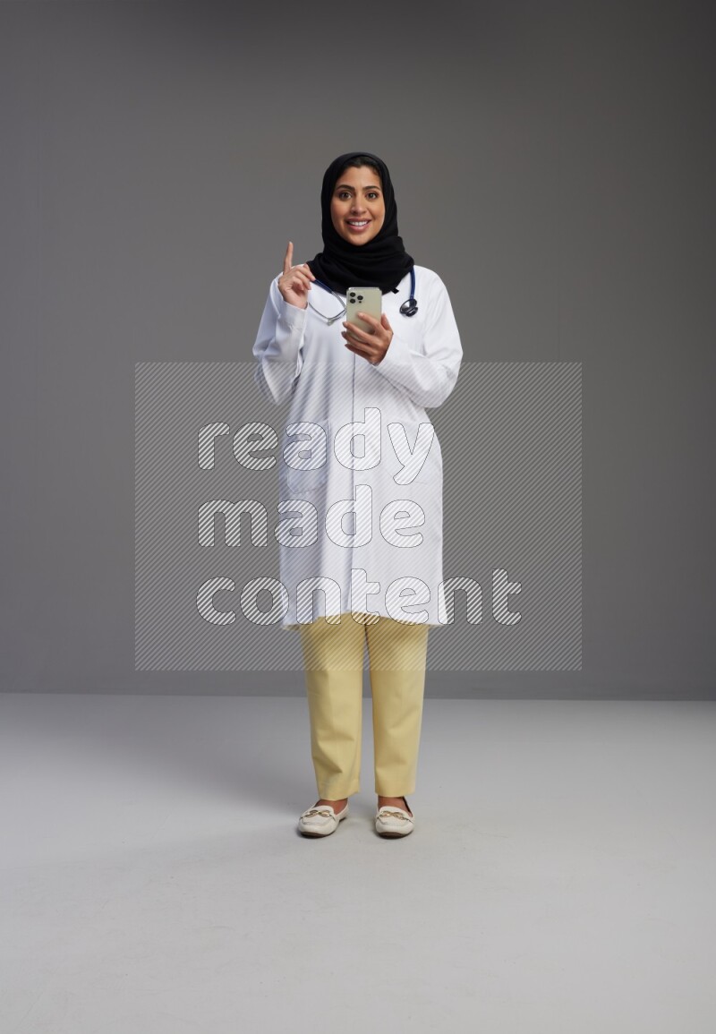 Saudi woman wearing lab coat with stethoscope standing texting on phone on Gray background