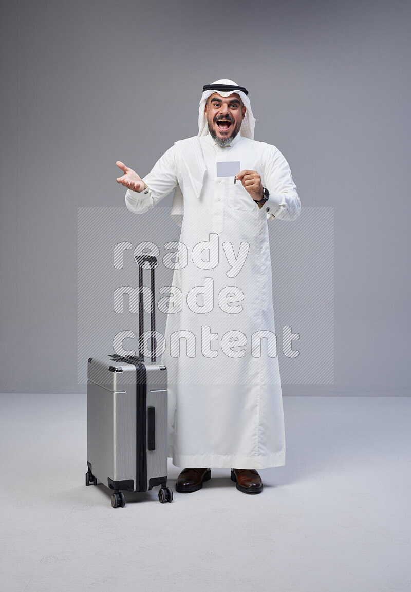 Saudi man wearing Thob and white Shomag standing holding Travel bag and ATM card on Gray background