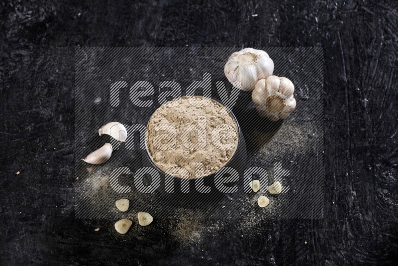 A black pottery bowl full of garlic powder with garlic cloves and bulb beside it on a textured black flooring in different angles